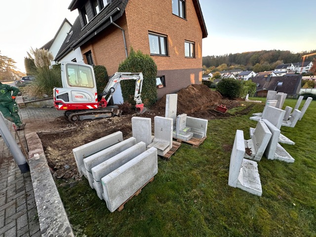 Ein Minibagger arbeitet neben einem Ziegelhaus, Betonplatten sind auf dem Rasen gestapelt.