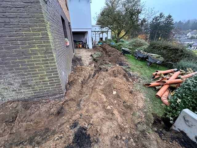Baustelle neben Ziegelgebäude mit ausgehobenem Graben und orangefarbenen Rohren im Vordergrund.