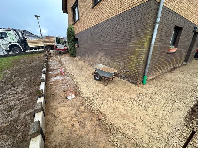 Baustelle neben einem Gebäude mit brauner Ziegelfassade, Schubkarre, Bagger und Lastwagen im Hintergrund.