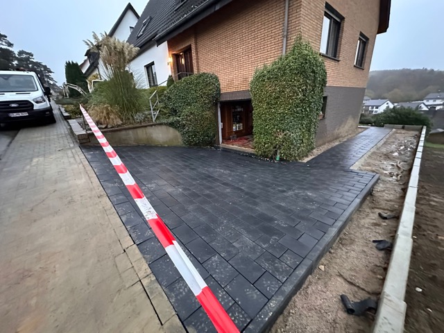 Zweistöckiges Ziegelhaus mit neuer, abgesperrter Pflastereinfahrt, umgeben von grünen Pflanzen.