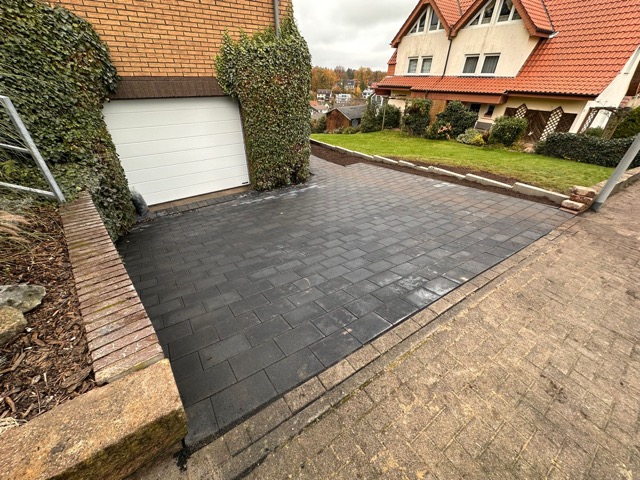 Eine frisch gepflasterte Einfahrt vor einem Haus mit Efeu, rotem Dach und einem kleinen Garten.