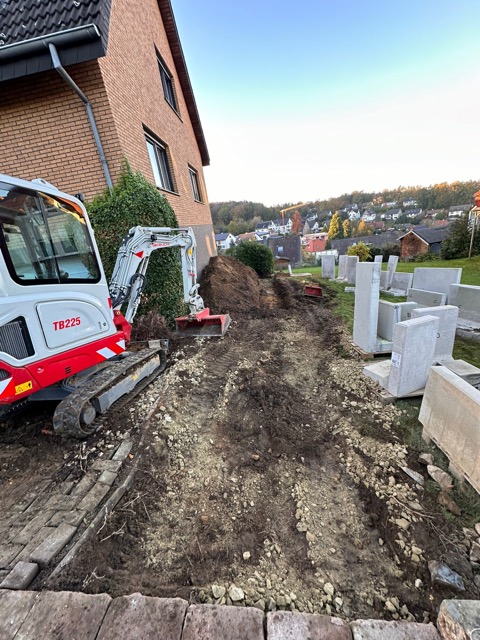Ein kleiner Bagger arbeitet neben einem Ziegelgebäude auf einer Baustelle mit Betonblöcken und Erdaushub.