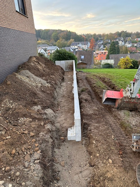 Baustelle neben einem Gebäude mit Ziegelmauer, Betonfundament und Bagger; ruhige, alltägliche Szene.
