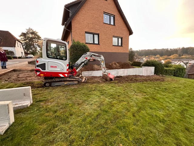 Ein Minibagger arbeitet auf einer Rasenfläche vor einem Backsteinhaus. Betonblöcke sind aufgestellt.