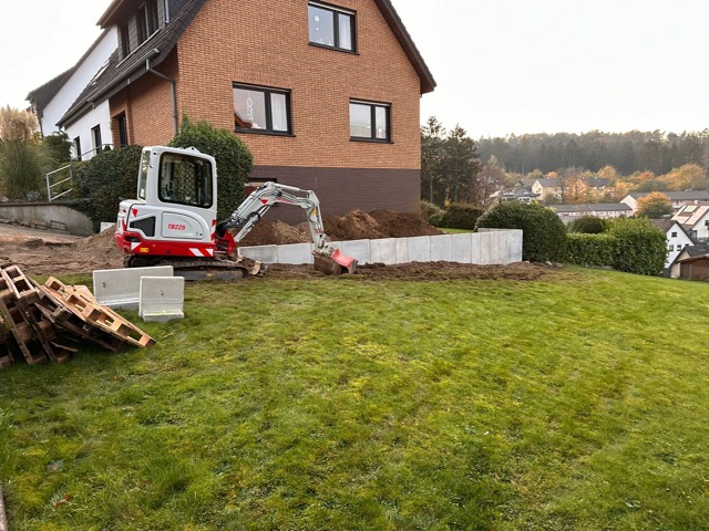 Eine Baustelle vor einem Ziegelhaus mit Minibagger, Betonplatten und Holzpaletten im Garten.