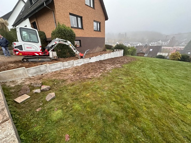 Baustelle neben Wohnhaus mit Minibagger und Betonblöcken, bewölkter Himmel, ruhige Arbeitsatmosphäre.
