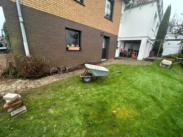Ein ruhiger Garten mit Schubkarre im Vordergrund und einem Ziegelhaus im Hintergrund.