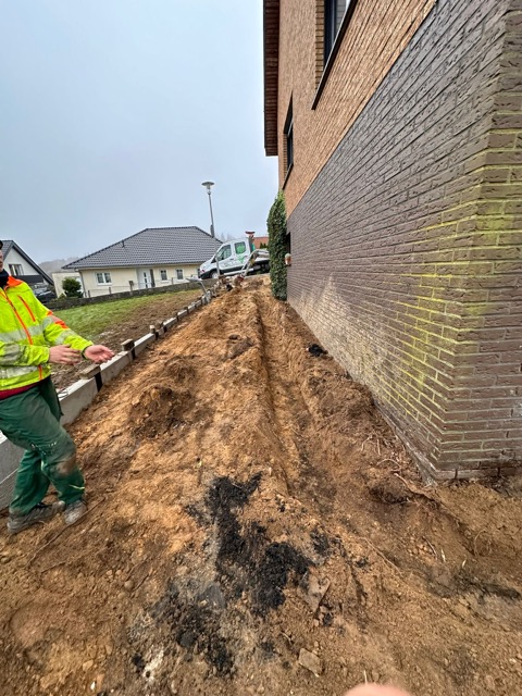 Ein Arbeiter in Warnkleidung steht auf einer Baustelle neben einem Ziegelgebäude unter grauem Himmel.