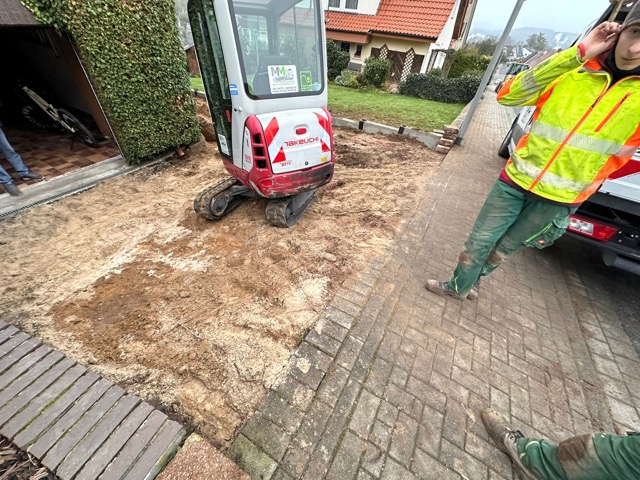 Ein Bagger steht still auf einer Baustelle; eine Person in Warnkleidung telefoniert daneben.