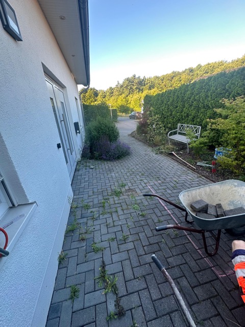 Gepflasterter Weg neben weißem Haus, Schubkarre mit Steinen, umgeben von grünem Garten.