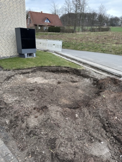 Baustelle vor einem Gebäude mit aufgewühlter Erde, einem weißen Zaun und einem schwarzen Objekt.