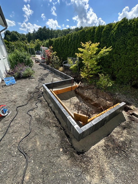 Eine Baustelle im Garten mit Betonfundament, Werkzeugen und Pflanzen im Hintergrund an einem sonnigen Tag.