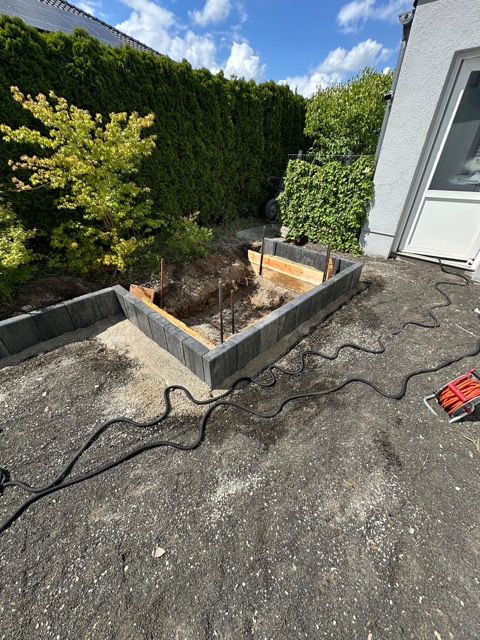 Baustelle im Garten mit halbfertiger Betonmauer, Metallstangen und umgebender Vegetation.