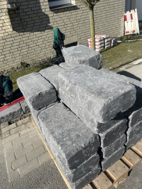 Ein Stapel grauer Pflastersteine auf einer Palette vor einer Ziegelmauer, umgeben von Baumaterialien.