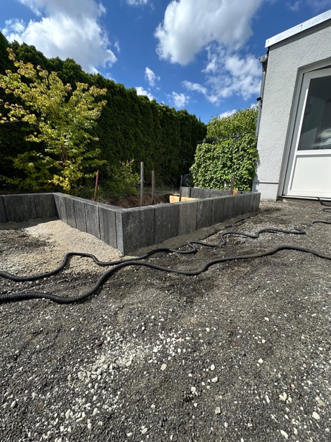 Baustelle im Garten mit Hochbeet, Schläuchen und Pflanzen neben einem Gebäude mit weißer Fassade.