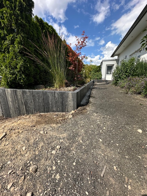 Moderner Garten mit Kiesweg, Holzmauer, grünen Pflanzen, rotem Baum und weißem Gebäude im Hintergrund.