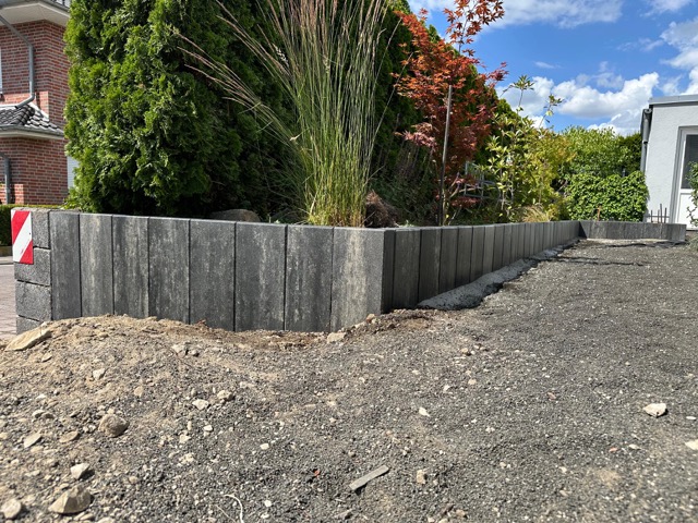 Garten mit grauer Steinmauer, Pflanzen und Baum mit roten Blättern, Haus im Hintergrund.