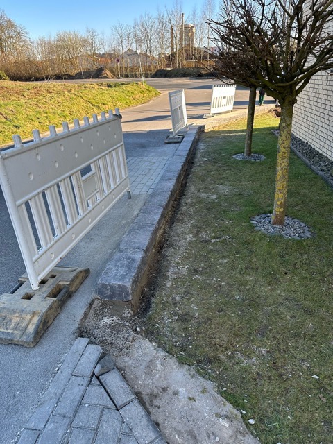 Baustelle mit weißen Absperrungen, kahlem Baum und gepflastertem Gehweg neben einer Ziegelmauer.