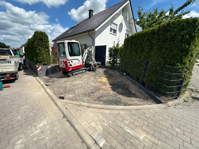 Ein Minibagger steht vor einem weißen Einfamilienhaus auf einer Baustelle, umgeben von Hecke und Zaun.