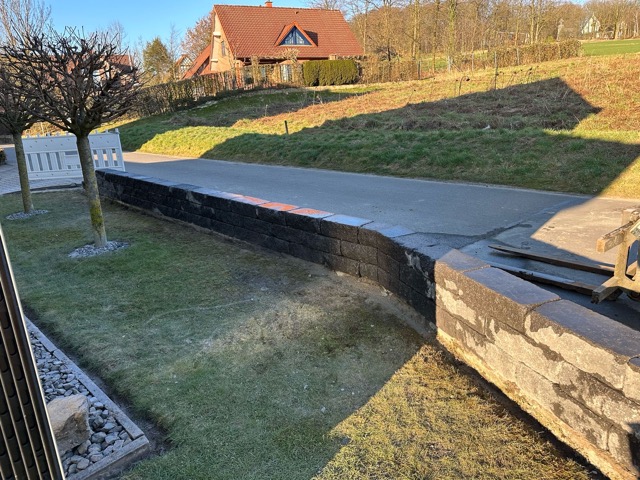 Gepflasterte Straße mit dunkler Steinmauer, grünem Garten und Häusern mit roten Dächern im Hintergrund.