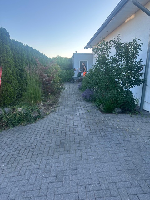 Zwei Personen schieben eine Schubkarre auf einem gepflasterten Weg neben einem weißen Gebäude.