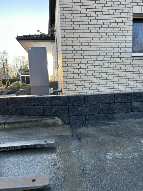 Moderne Gebäudeecke aus hellen Backsteinen, dunkle Mauer, feuchter Asphalt, klarer blauer Himmel.