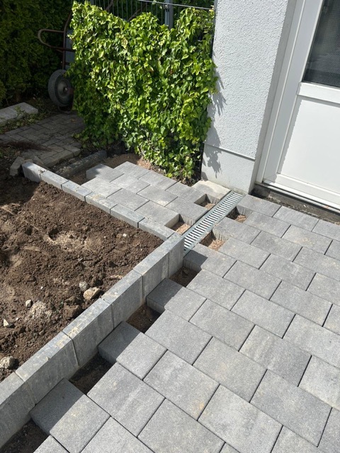 Neu gepflasterte Fläche mit grauen Steinen, Entwässerungsrinne und angrenzender Erdfläche neben Gebäude.
