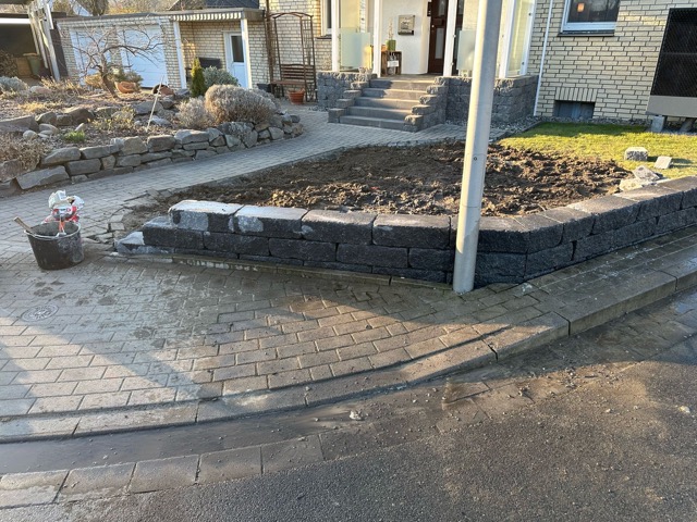 Ein gepflegter Vorgarten mit einem Beet aus grauen Steinblöcken, daneben ein Gehweg und ein Haus.