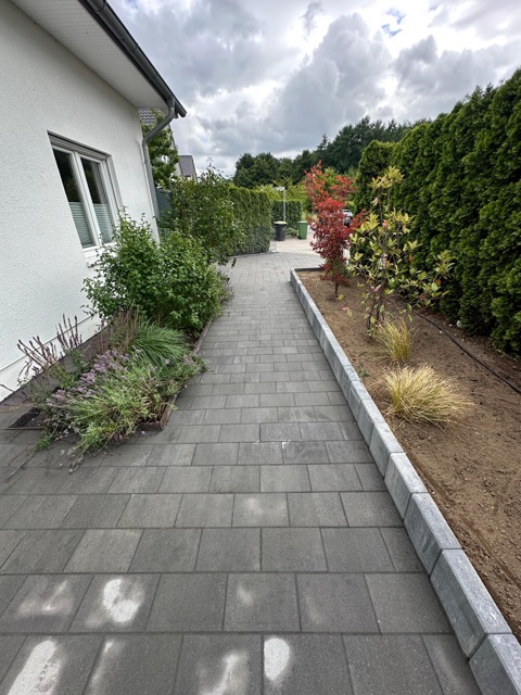 Ein gepflasterter Gartenweg führt an einem weißen Haus vorbei, flankiert von Pflanzen und einem roten Strauch.