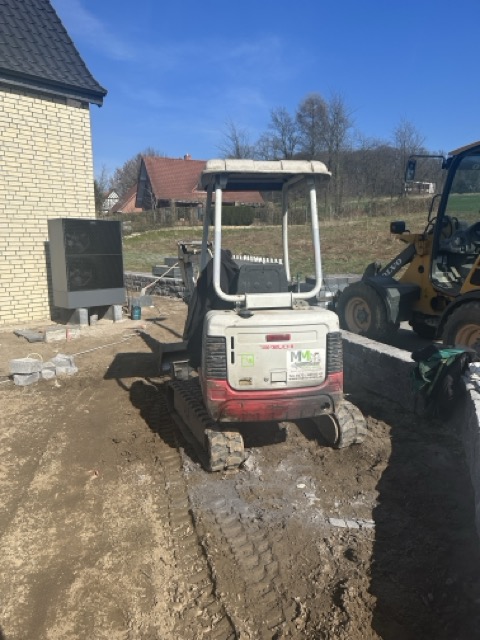 Ein kleiner Bagger und ein Radlader stehen auf einer ruhigen Baustelle vor einem gelben Backsteingebäude.