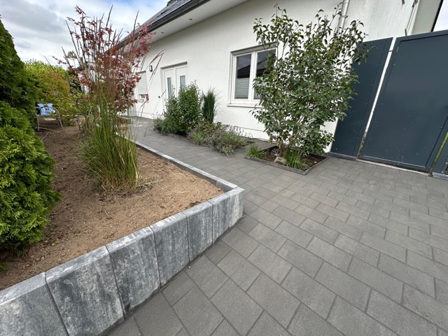 Moderner Hauseingang mit weißer Fassade, grauem Pflasterweg und grünem Hochbeet, ruhig und gepflegt.