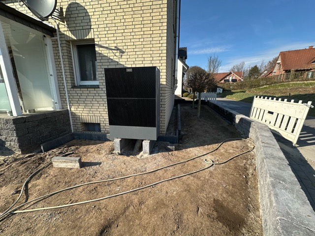 Gelbes Backsteinhaus mit montierter schwarzer Box und Satellitenschüssel, erdiger Boden mit Kabeln.