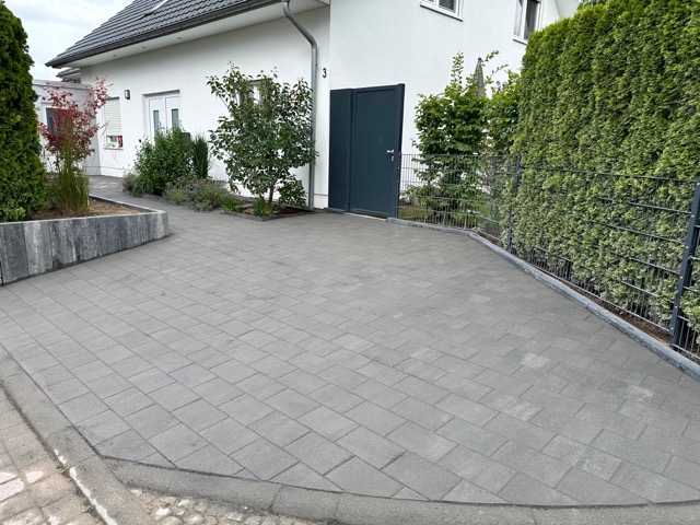 Moderner, weißer Hausvorhof mit grauen Pflastersteinen, grüner Vegetation und einem Metallzaun.