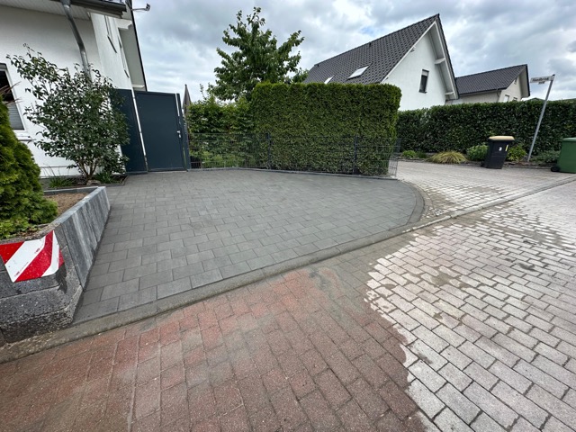 Moderne Einfahrt mit grauen Pflastersteinen, umgeben von Pflanzen, Hecken und weißen Wohnhäusern.