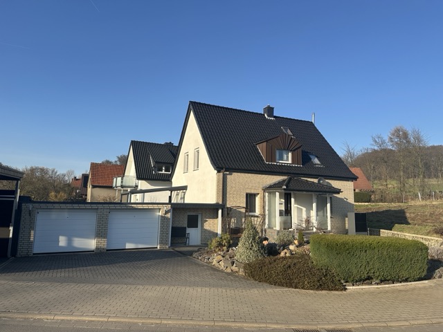 Ein zweistöckiges Einfamilienhaus mit schwarzem Dach, Doppelgarage und gepflegtem Vorgarten bei Sonnenschein.