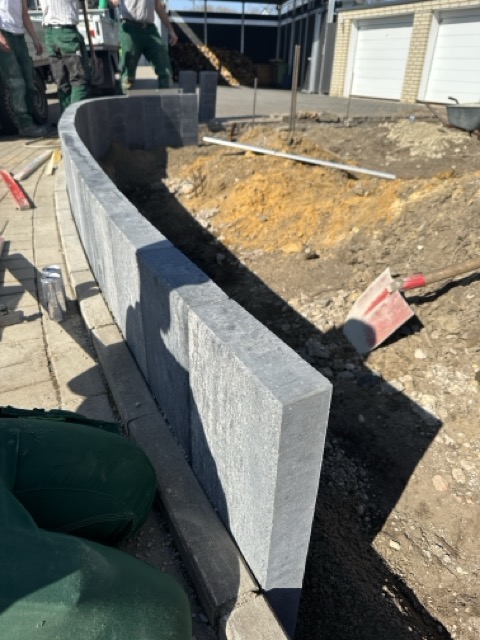 Baustelle mit Arbeitern, die eine geschwungene Mauer aus grauen Steinblöcken errichten.