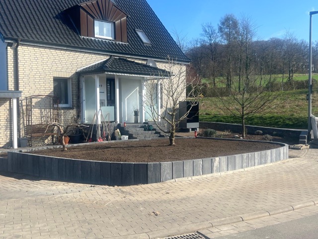 Ein Einfamilienhaus mit hellem Klinker, dunklem Dach und frisch bearbeitetem Vorgarten unter blauem Himmel.