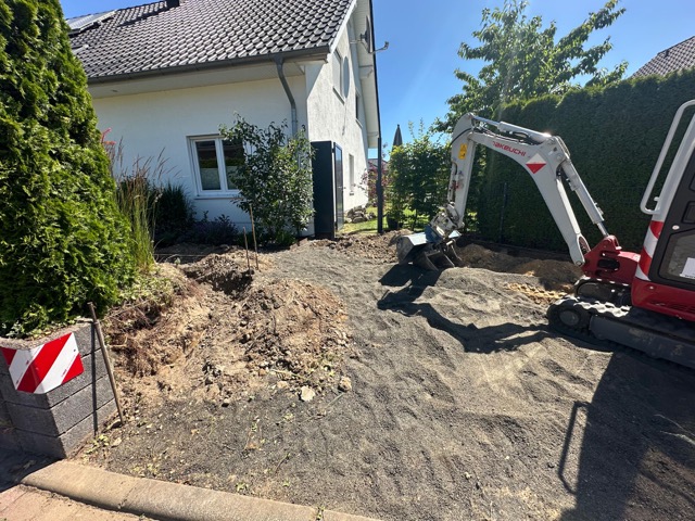 Ein Minibagger arbeitet in einem Garten neben einem weißen Haus, umgeben von Pflanzen und Bäumen.