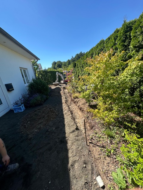 Ein weißes Haus mit Gartenarbeiten; ein Bagger gräbt neben Pflanzen und Hecke bei sonnigem Wetter.