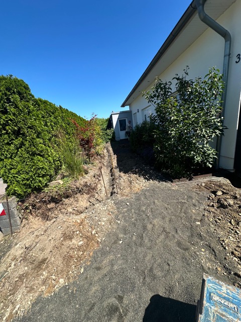 Ein schmaler Weg neben einem Haus, umgeben von Sträuchern, mit sichtbaren Gartenarbeiten.