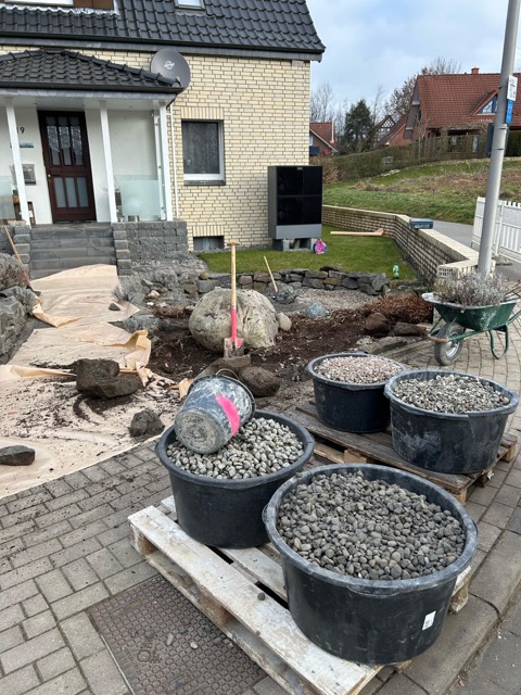 Baustelle vor einem gelben Backsteinhaus mit Kübeln, Schubkarre, Felsbrocken und Gartengeräten.
