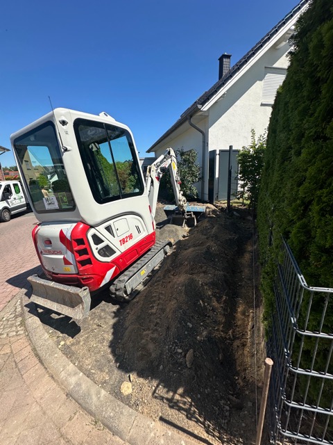 Ein kleiner Bagger steht auf Pflastersteinen neben einem frisch ausgehobenen Graben vor einem Wohnhaus.