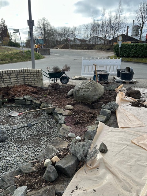 Eine Baustelle mit Schubkarre, Steinen und Pflanzen zeigt laufende Gartenarbeiten im Freien.