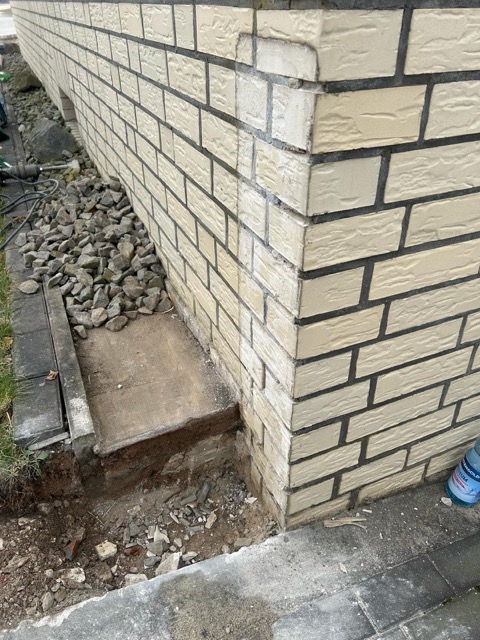 Ecke eines Gebäudes mit heller Klinkerfassade, Kies und Betonfundament; Wasserflasche deutet auf Arbeiten hin.