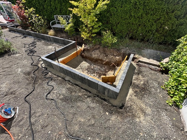 Baustelle im Garten mit Betonfundament, Holzverschalungen, Gartenschlauch und grüner Hecke im Hintergrund.