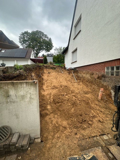 Baustelle in Wohngebiet mit frisch ausgehobenem Hang, Haus mit weißer Fassade und roten Klinkern.