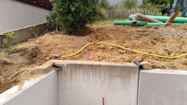 Baustelle mit Betonwand, Erdhügel, gelbem Schlauch, grünen Rohren und einer Pflanze im Freien.