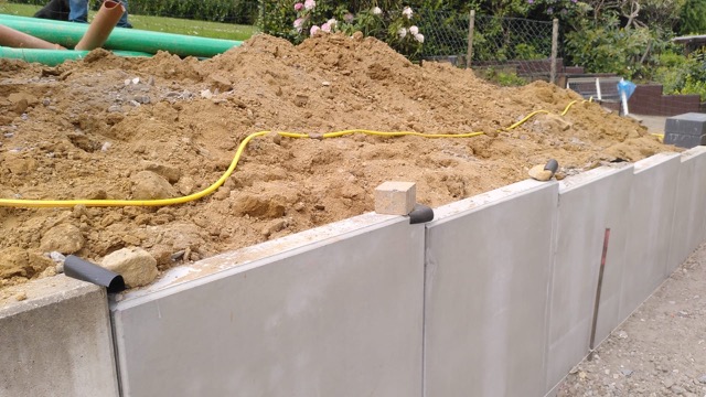 Eine Baustelle mit Betonmauer, Erdhaufen, gelbem Schlauch und Baumaterialien im Hintergrund.