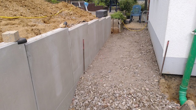 Baustelle mit Betonmauer, Schotterweg, weißem Gebäude und Werkzeugen; ruhige, arbeitsame Atmosphäre.
