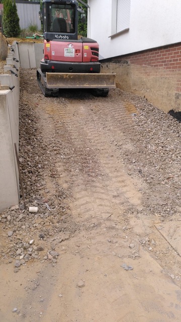 Ein kleiner roter Bagger steht still auf einer Schotterfläche zwischen Betonwänden auf einer Baustelle.